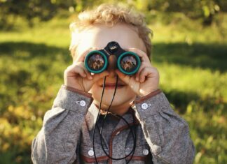 Young blonde boy looking through binoculars during lifestyle portrait session in apple orchard at golden hour.