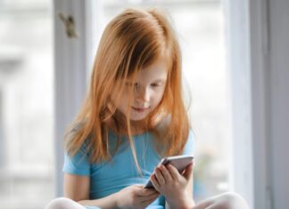 Calm small ginger girl sitting on table and using smartphone in light living room