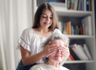 Cheerful teenager playing with grandmother guess who game while making surprise in light living room