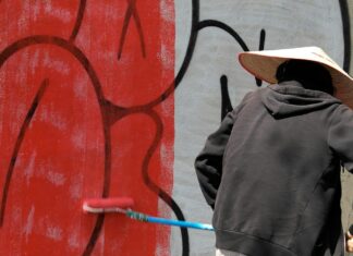 A young man with a hat painting a dirty wall with a roller with red paint