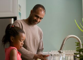 This African-American father was shown in the process of teaching his young daughter how to properly wash her hands at their kitchen sink, briskly rubbing her soapy hands together under fresh running tap water, in order to remove germs, and contaminants, thereby, reducing the spread of pathogens, and the ingestion of environmental chemicals or toxins. Children are taught to recite the Happy Birthday song, during hand washing, allotting enough time to completely clean their hands.